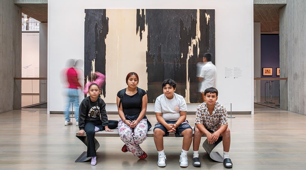 Four children sit on a bench in an art museum gallery in front of a large abstract painting